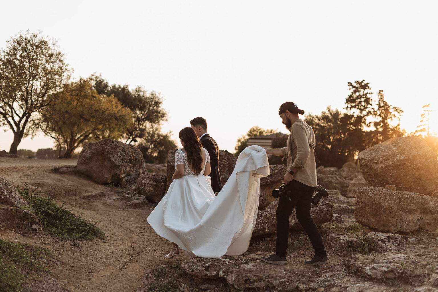 Il fotografo aiuta la sposa con lo strascico mentre gli sposi camminano tra gli ulivi al tramonto