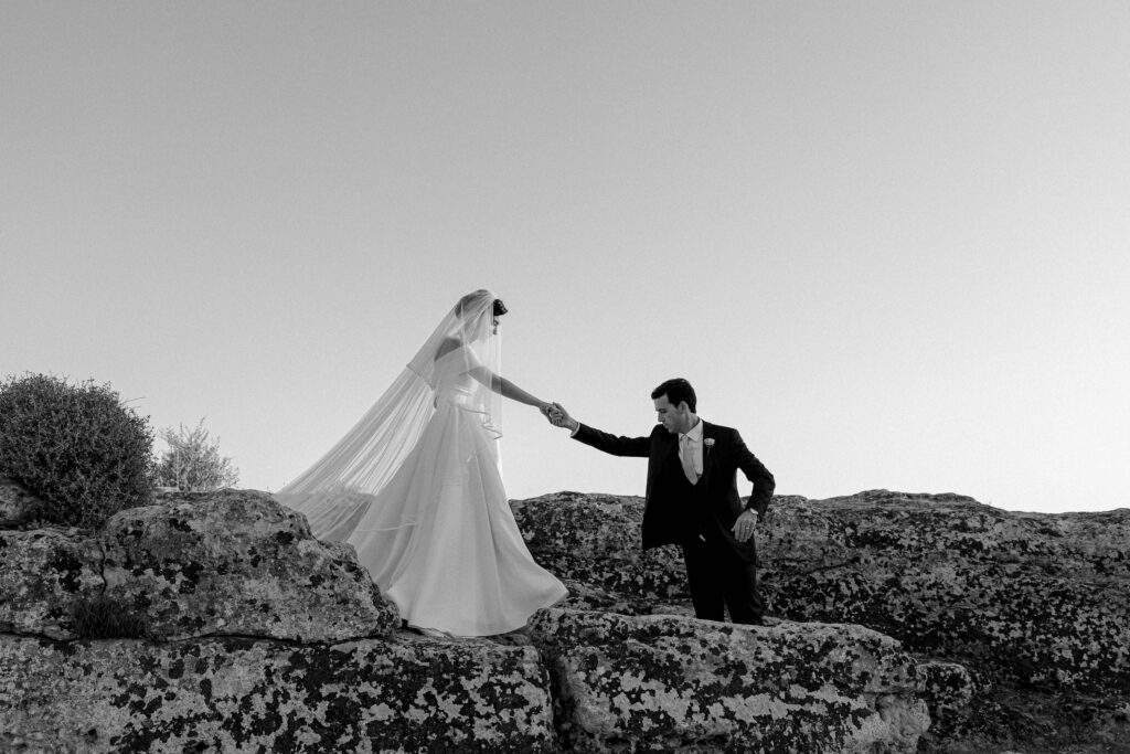 Sposi che si tengono per mano sulle rocce, il velo della sposa al vento – matrimonio in Sicilia in bianco e nero