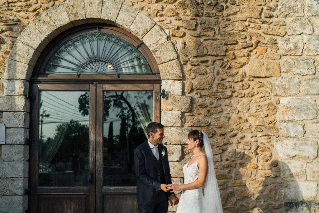 Lia e Giorgio si tengono per mano davanti a un muro di pietra con un portone ad arco